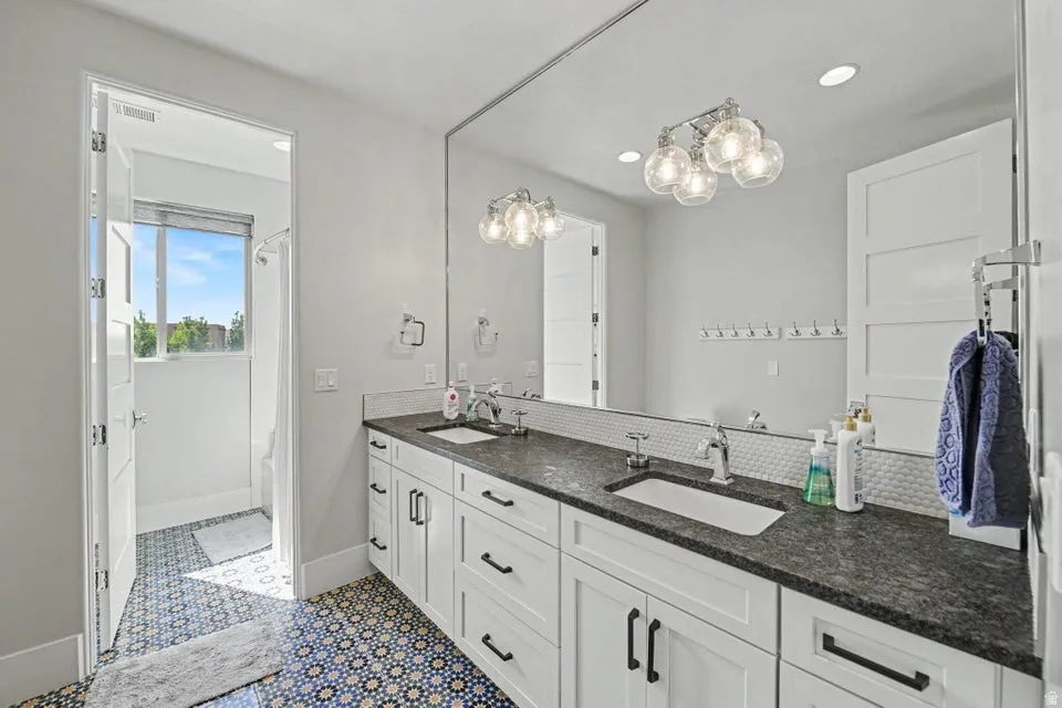 Double vanity with dark stone countertop, white shaker cabinetry, large wall-mounted mirror, and decorative tile flooring
