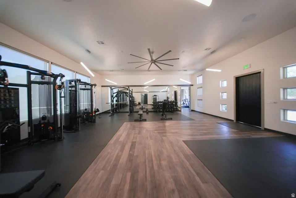 Spacious exercise room featuring wood-finish flooring, rubber matting, multiple windows with roller shades, and a large ceiling fan