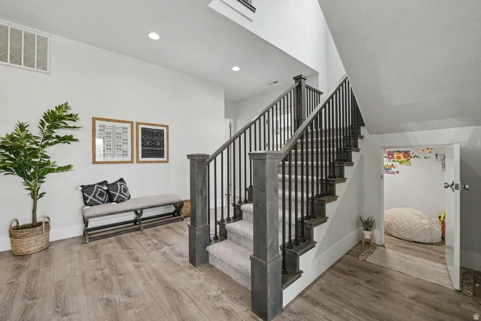 Wood-finish flooring, staircase with dark wood newel posts, black iron balusters, and carpeted treads, recessed ceiling lighting
