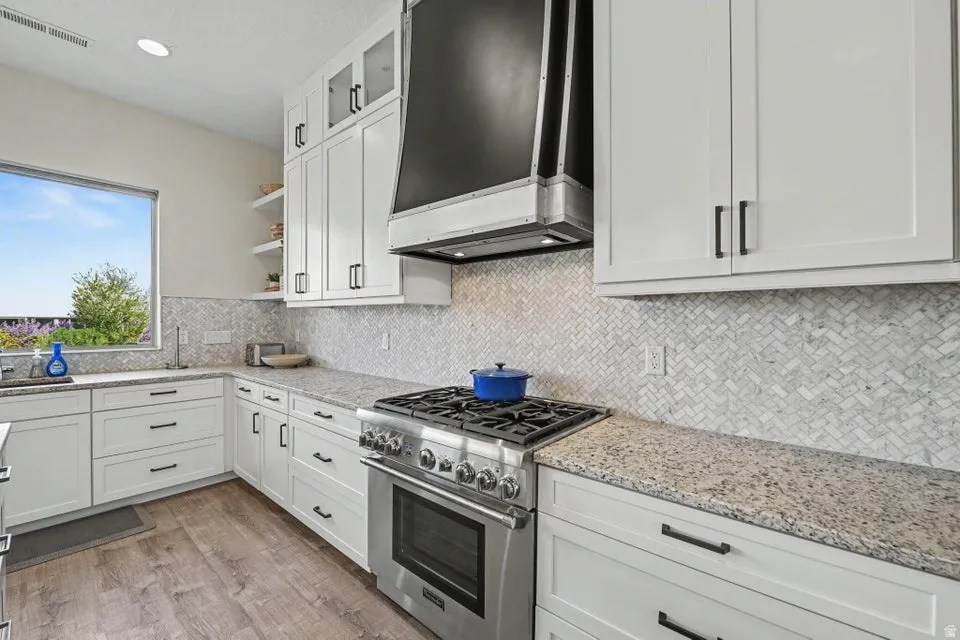 White shaker cabinetry, herringbone tile backsplash, stainless steel range with hood, stone-finish countertops, and wood-finish flooring