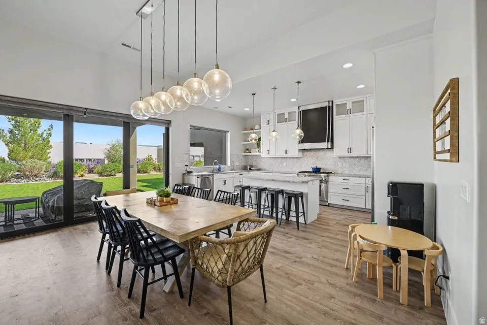 Expansive sliding glass doors, wood-finish flooring, a contemporary linear chandelier, white shaker-style cabinetry, and a kitchen island with bar seating