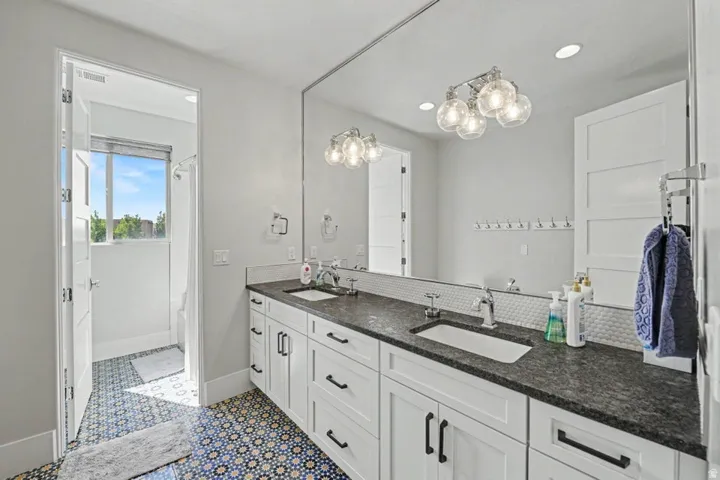 Double vanity with dark stone countertop, white shaker cabinetry, large wall-mounted mirror, and decorative tile flooring