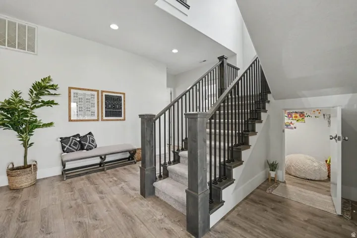 Wood-finish flooring, staircase with dark wood newel posts, black iron balusters, and carpeted treads, recessed ceiling lighting