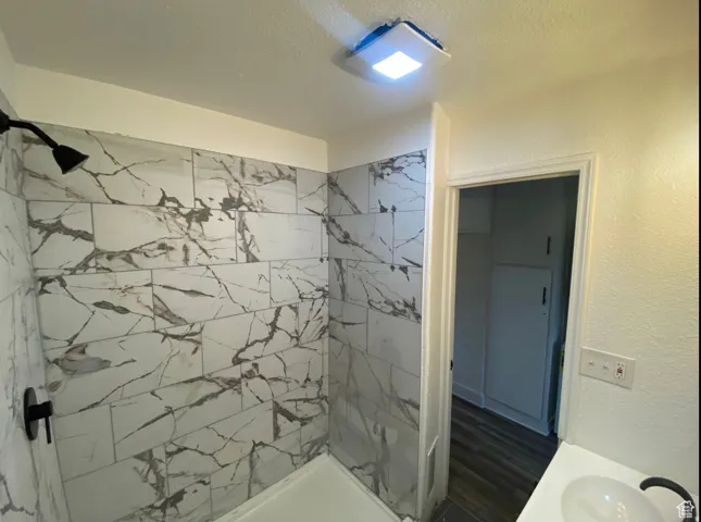 Shower with white and gray veined tile surround, matte black shower fixtures, a white porcelain sink, and wood-finish flooring