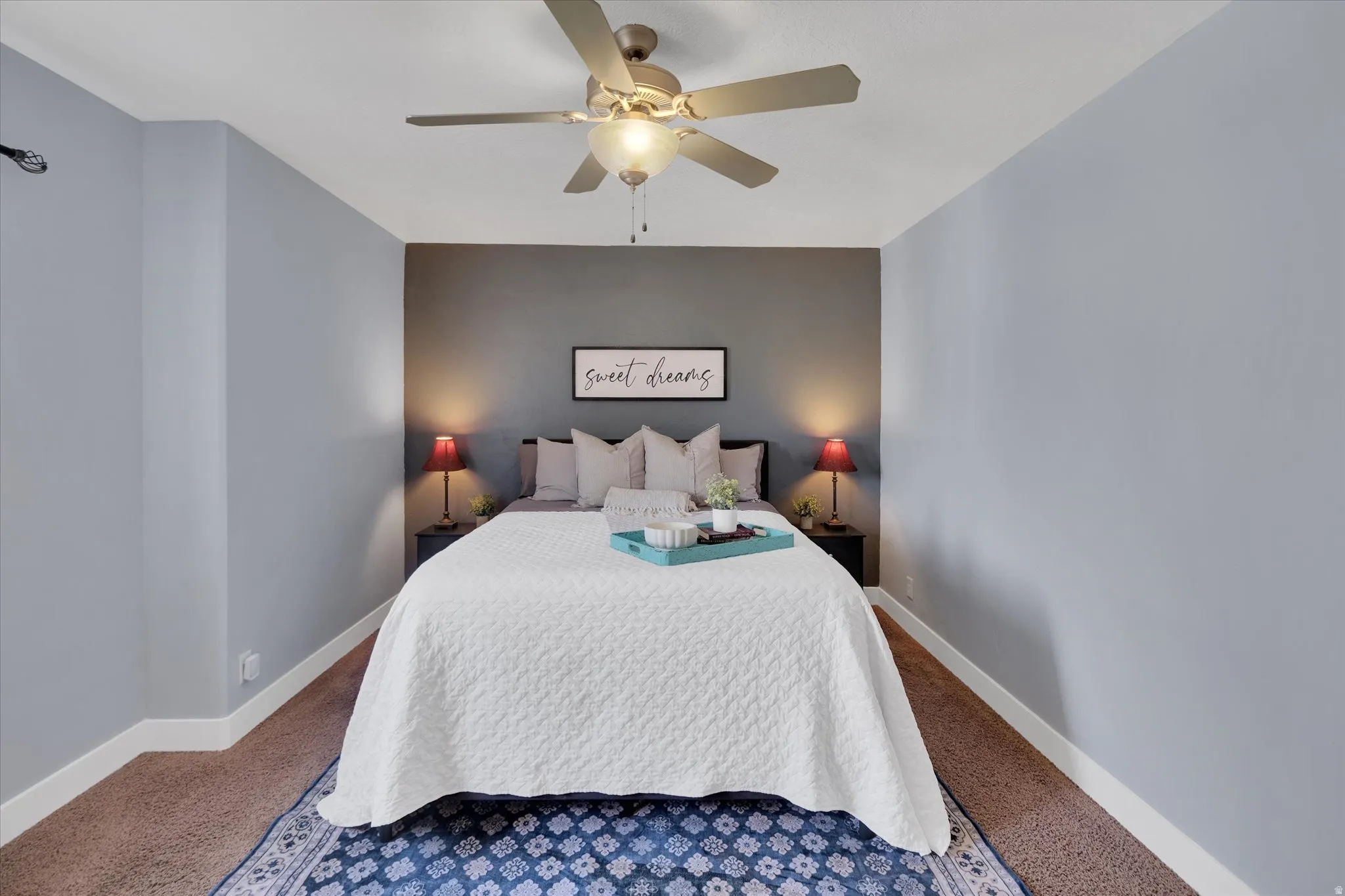 Bedroom featuring soft blue-gray walls, warm bedside lighting with table lamps, a ceiling fan with integrated light fixture, and wood-finish flooring complemented by a patterned area rug