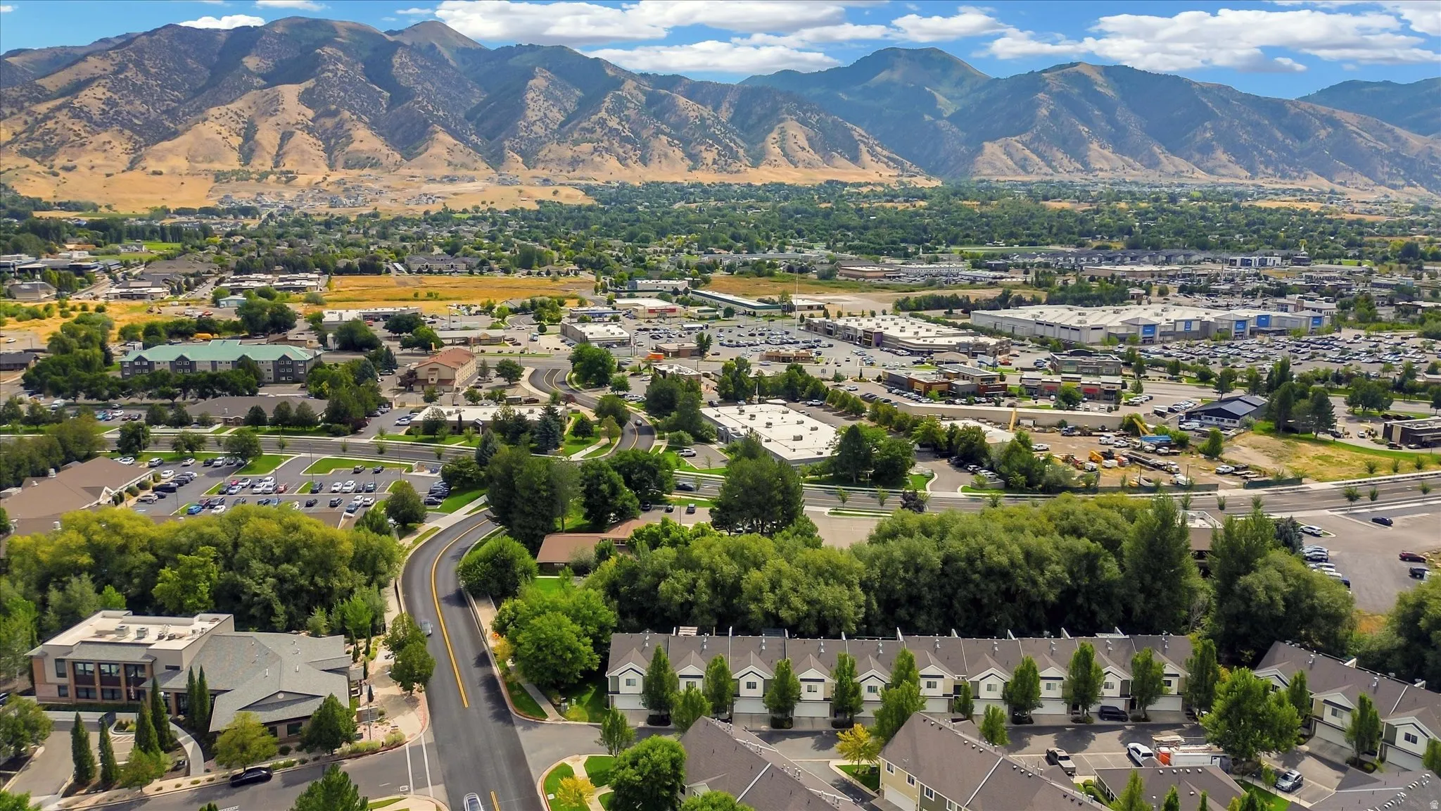 Aerial view showcasing a residential community nestled against dramatic mountain backdrop, featuring tree-lined streets, established landscaping throughout the neighborhood, and proximity to commercial corridors and retail developments visible in the surr
