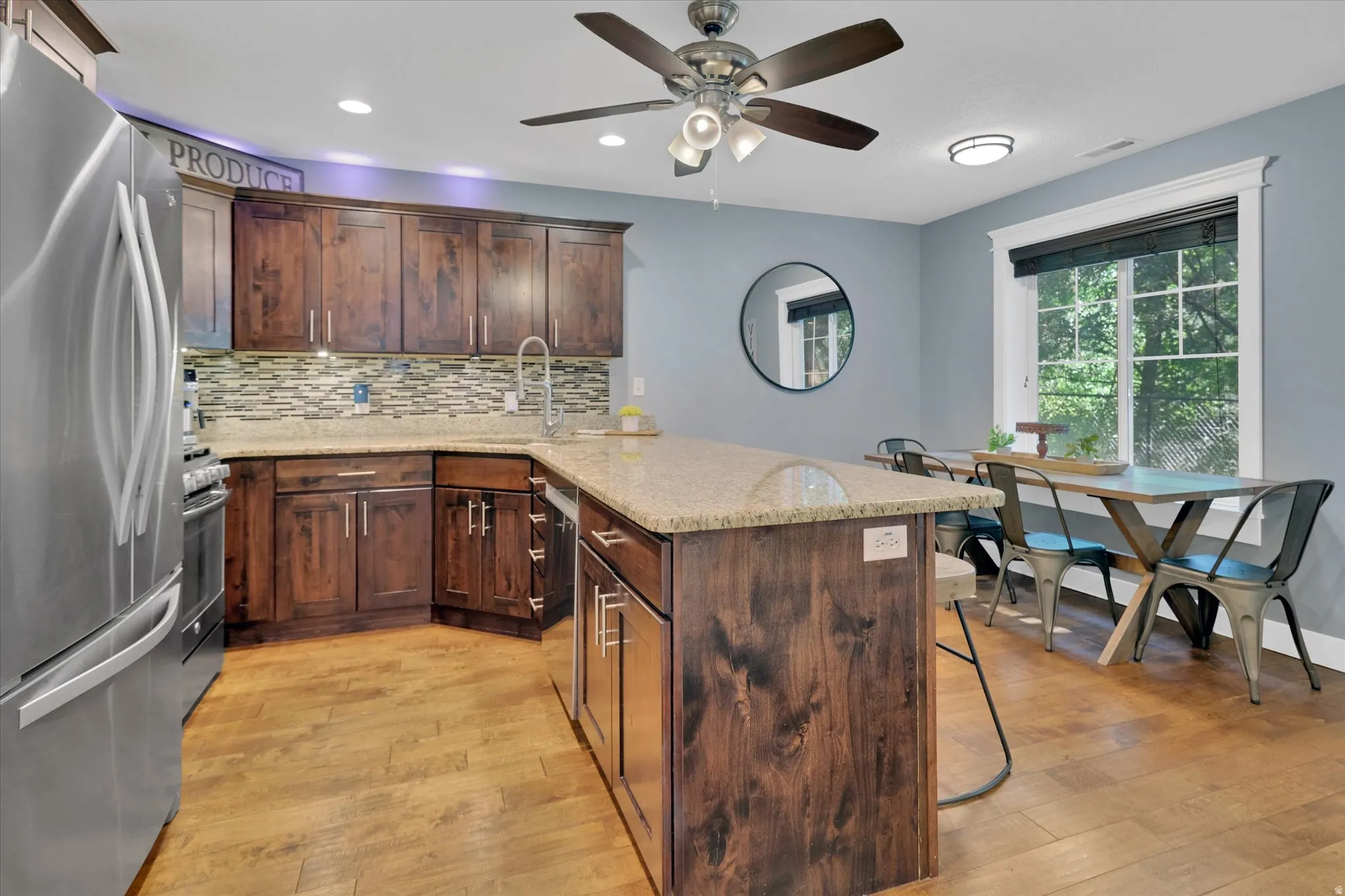 Kitchen featuring wood-finish flooring, dark wood cabinetry, a stainless steel refrigerator, stone countertops, and a tile backsplash