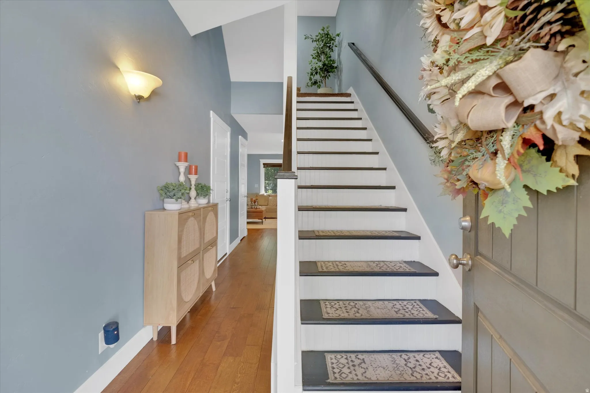 Elegant foyer featuring hardwood flooring, a staircase with carpeted treads and white railings, soft wall sconce lighting, and neutral wall finishes