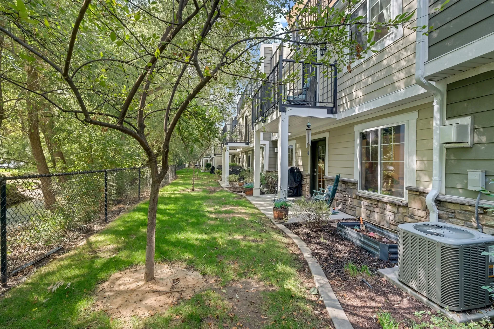 Exterior siding with stone detailing, an upper-level balcony with metal railing, a ground-level patio, and a grassy area bordered by mature trees