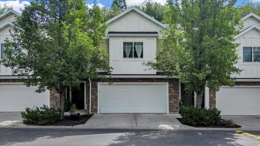 Two-story facade with stone and siding accents, prominent white garage doors, and mature trees providing natural shading