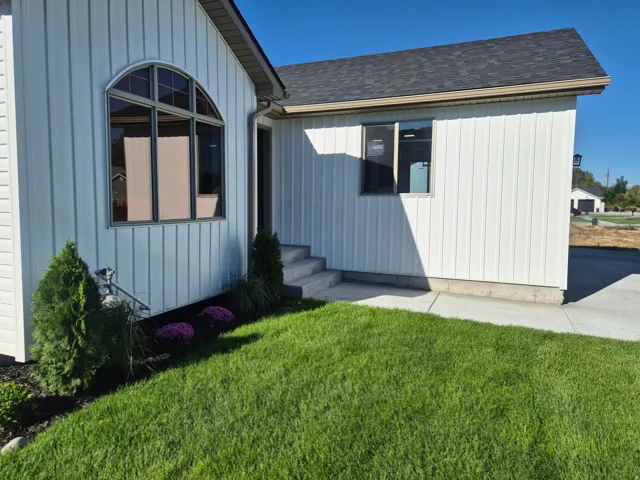 Arched window with dark framing, white vertical siding, dark asphalt shingle roof, concrete walkway, and manicured lawn