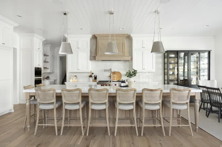 Expansive kitchen island with a light-toned countertop, complete with white perimeter cabinetry, wood-finish flooring, and a unique wood-toned range hood