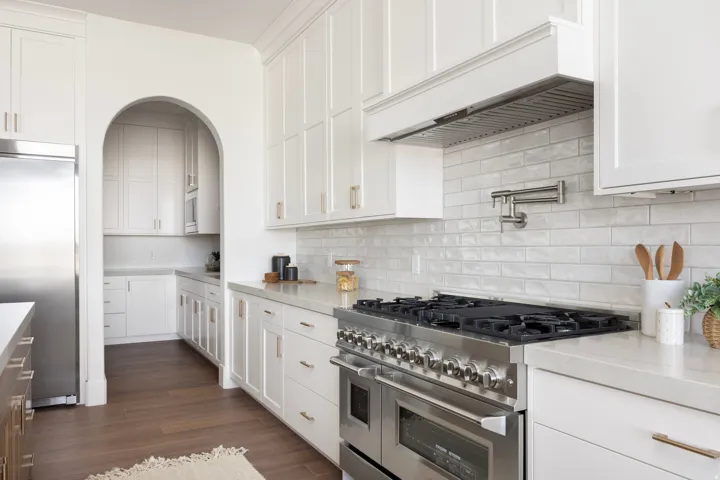 Gourmet kitchen featuring a stainless steel professional-grade range, white shaker cabinetry, light-toned countertops, a full-height tile backsplash, and a convenient pot filler