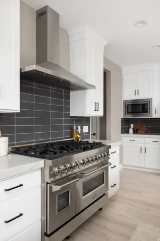 Stainless steel range with multiple burners, stainless steel range hood, white cabinetry with black hardware, white countertops, and dark tile backsplash