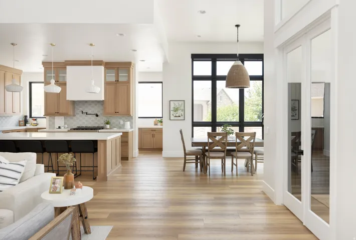 Open-concept living space featuring warm wood-finish flooring throughout, custom wood cabinetry with integrated appliances, quartz countertops, pendant lighting, and expansive black-framed glass doors and windows flooding the space with natural light