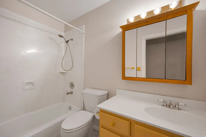White bathtub with shower, toilet, wood-finish vanity with an integrated sink, and a mirrored medicine cabinet with integrated lighting
