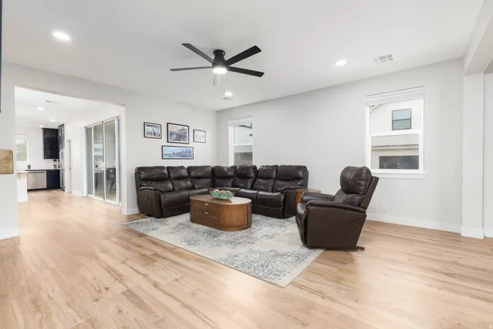 Spacious open-concept living area featuring wood-finish flooring throughout, a dark leather sectional sofa arrangement, ceiling fan with integrated lighting, recessed ceiling lights, and large windows providing natural illumination