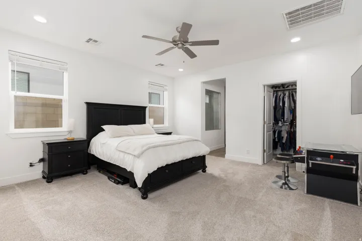Spacious primary bedroom featuring a black platform bed frame, neutral carpet flooring, ceiling fan with integrated lighting, large windows with cellular shades, and dedicated walk-in closet with organized storage system