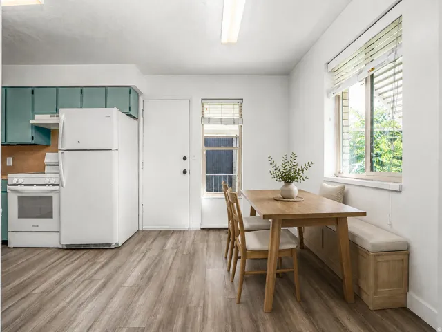 Wood-finish flooring throughout, complemented by a large window with blinds, teal cabinetry, and a built-in kitchen bench