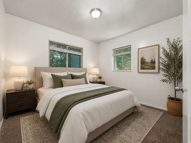 Serene primary bedroom featuring dual windows with green trim, neutral wall finishes, wood-finish nightstands with integrated lighting, natural fiber area rug, and decorative potted greenery