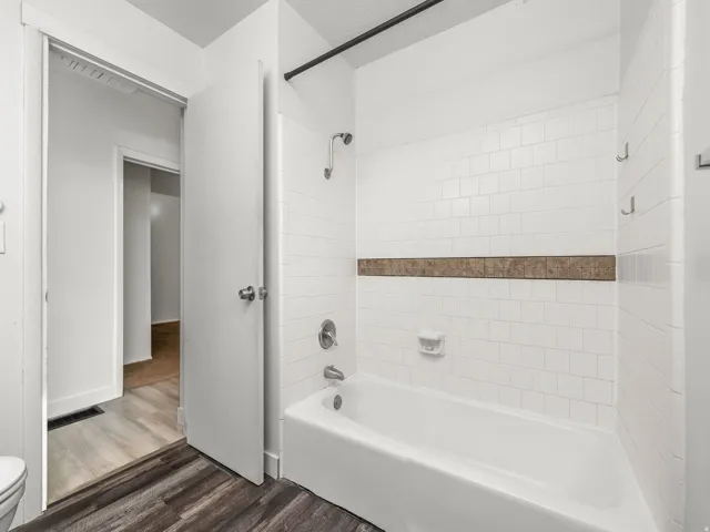 White subway tile shower surround with a decorative accent band, built-in bathtub, and dark wood-finish flooring