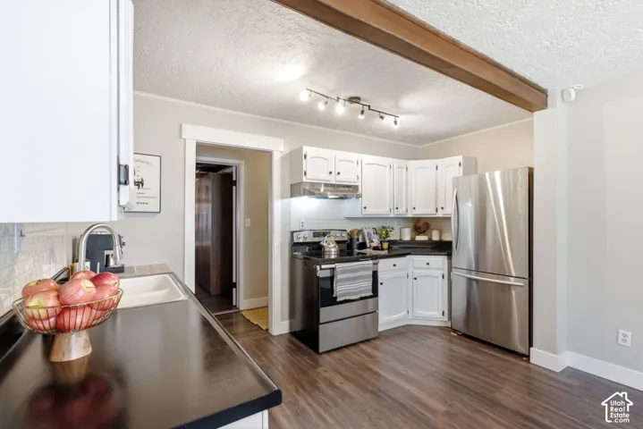 Kitchen featuring white cabinetry, black countertops, stainless steel appliances, wood-finish flooring, and exposed ceiling beam