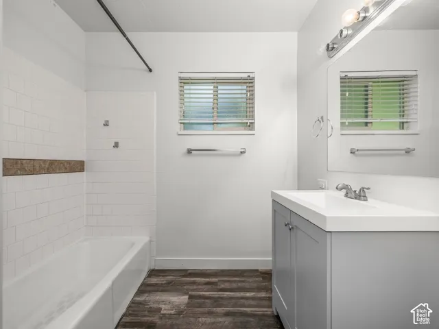 Spa-inspired primary bathroom featuring a ceramic tile surround with accent stripe, freestanding soaking tub, dual vanity with gray cabinetry and brushed nickel hardware, quartz countertop, and wood-finish plank flooring