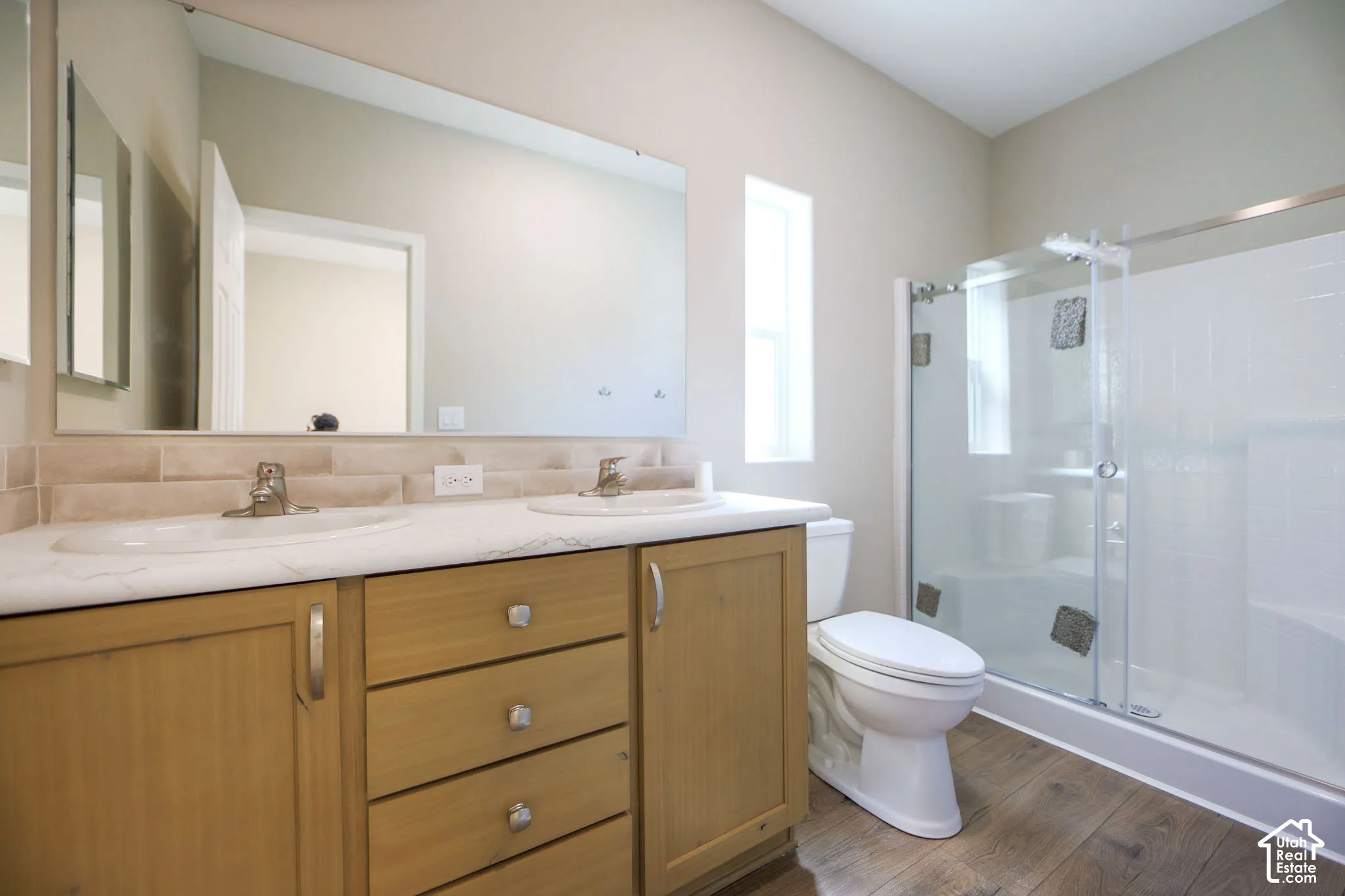 Dual basin vanity with wood-finish cabinetry, a large frameless mirror, and a tiled backsplash