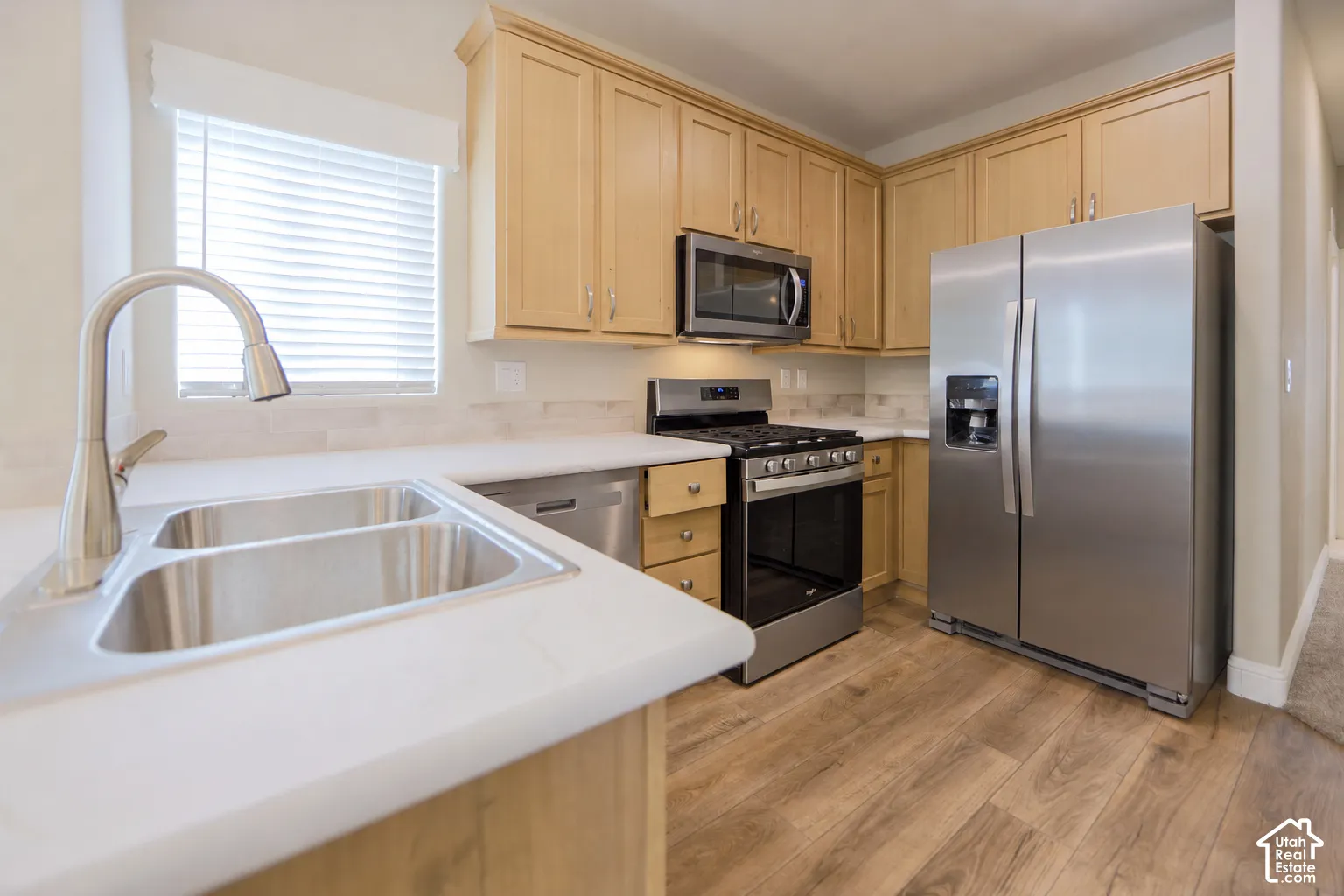 Contemporary kitchen featuring light wood cabinetry, stainless steel appliances, white countertops, a double basin sink with a gooseneck faucet, and wood-finish flooring