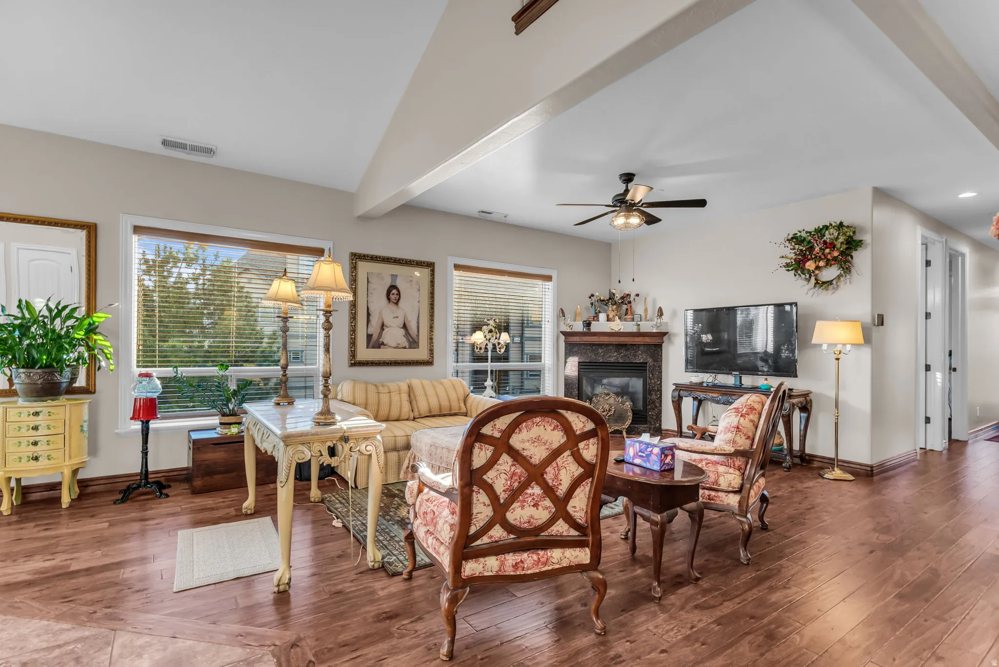 Vaulted ceiling with exposed beam, wood-finish flooring, multiple windows with blinds, built-in fireplace with dark stone surround, and a ceiling fan
