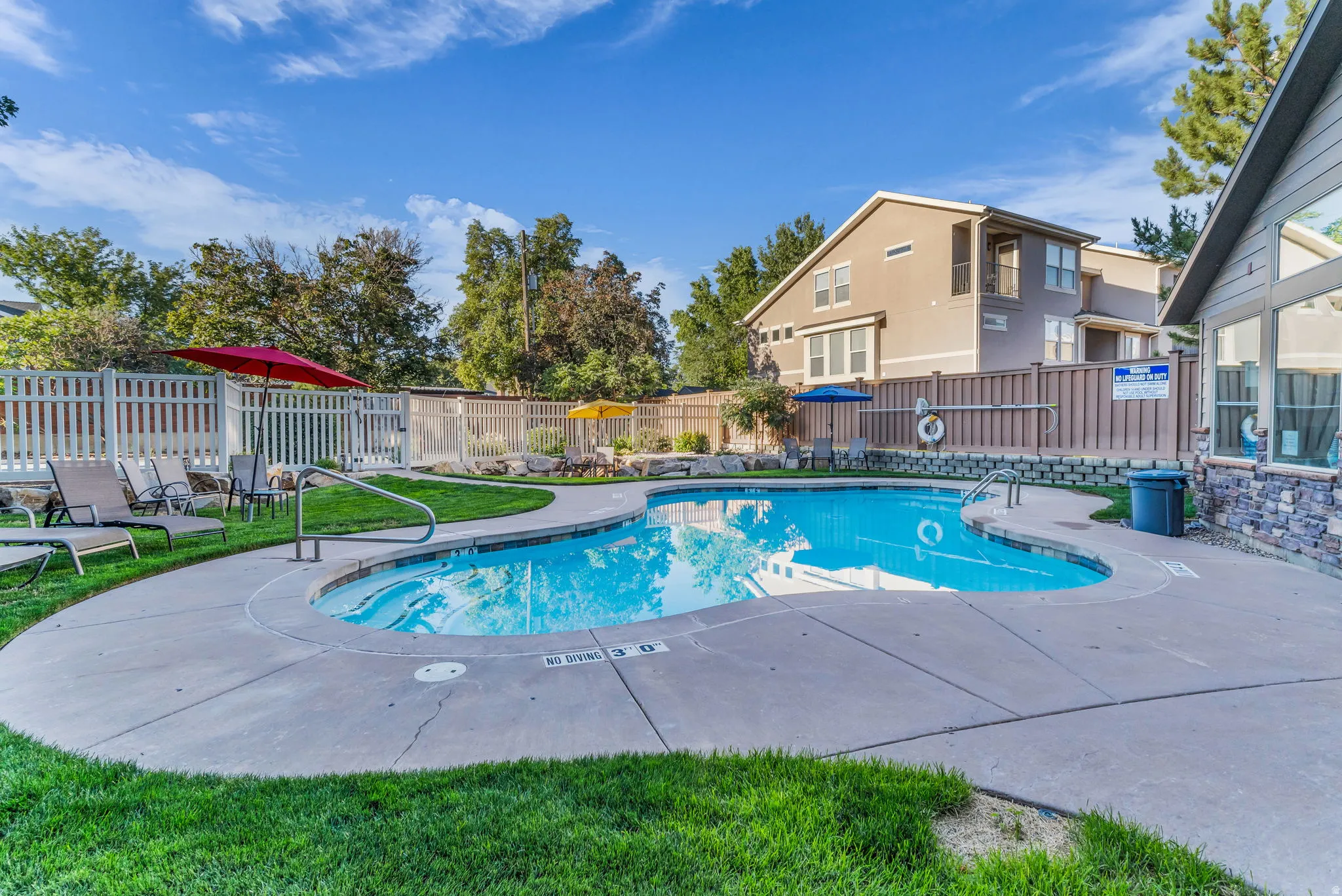 Outdoor pool with concrete decking, surrounding green space, and a white picket fence