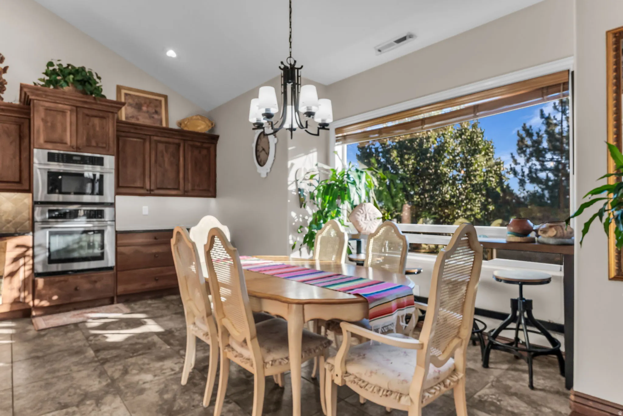 Vaulted ceiling, dark wood cabinetry, built-in stainless steel double oven, large picture window with wood blinds, and tile flooring