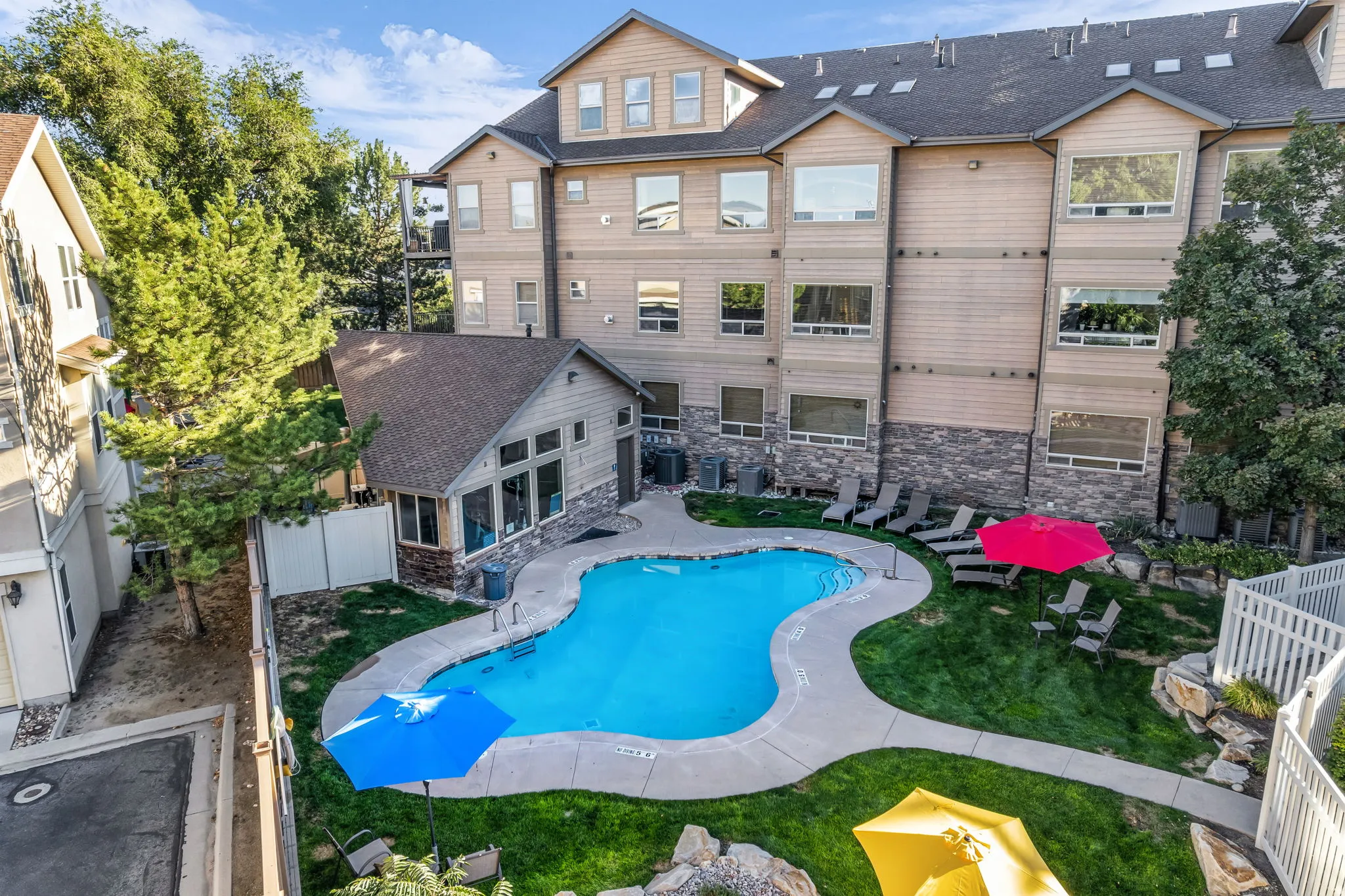 Curved swimming pool with surrounding concrete deck, adjacent landscaped lawn, and a stone-accented work out building with restrooms.