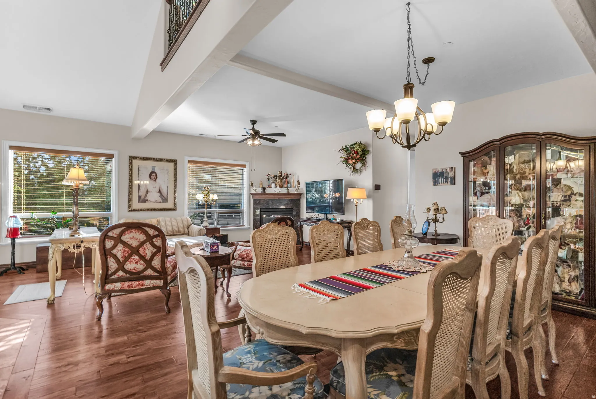 Vaulted ceiling with exposed beams, wood-finish flooring, two large windows, a built-in fireplace, and an overhead chandelier