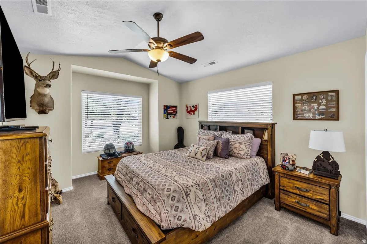 Vaulted ceiling, ceiling fan with integrated lighting, recessed window alcove, wood-finish bed frame with built-in storage, and wall-to-wall carpeting
