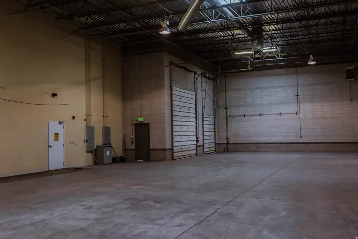 Concrete flooring, exposed structural ceiling, two roll-up doors, two pedestrian doors, and industrial lighting fixtures