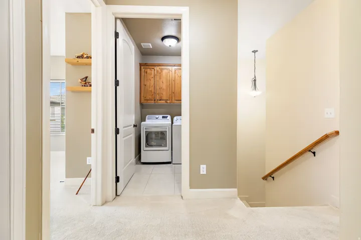 Carpeted hallway with a wood handrail, an overhead pendant light fixture, and a laundry area featuring wood cabinetry and tiled flooring