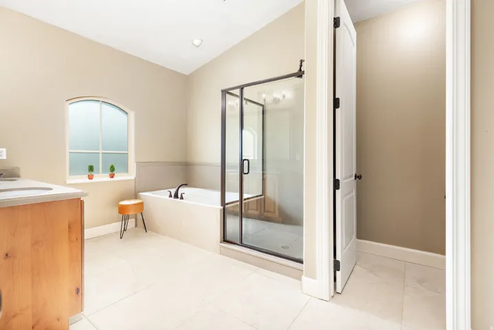 Arched window with frosted glass, built-in bathtub with dark fixtures, glass-enclosed shower, light wood-finish vanity with countertop, and light tile flooring