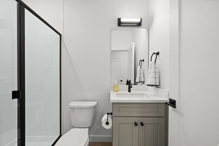 Shower with black framed glass enclosure, vanity with light gray shaker-style cabinetry, white countertop, rectangular undermount sink, and matte black faucet