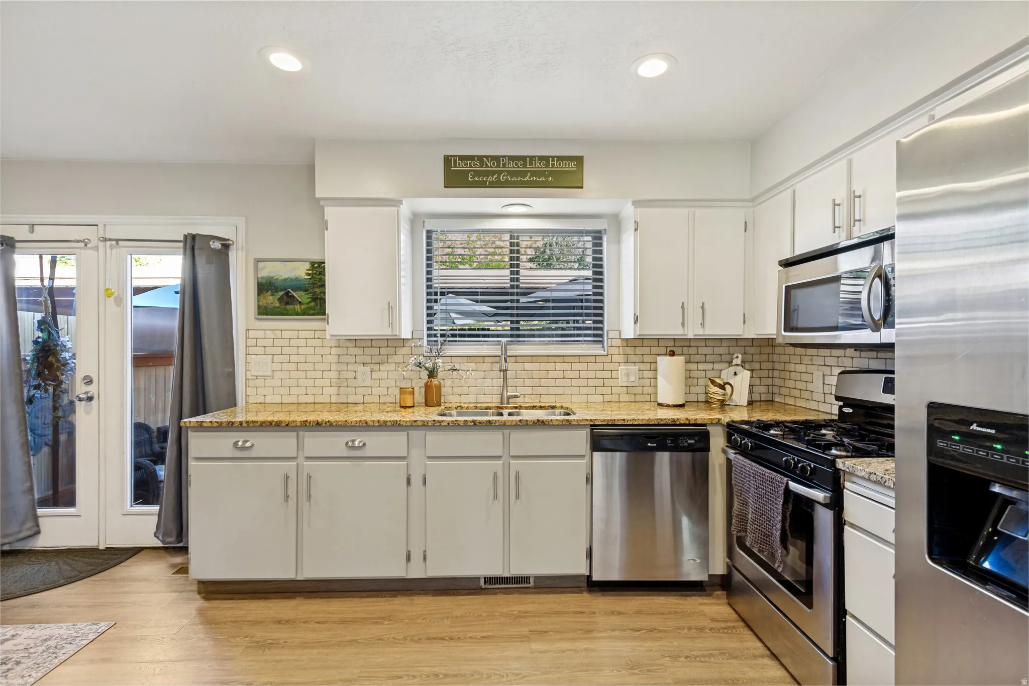 White cabinetry with brushed nickel hardware, stainless steel appliances, light-toned wood-finish flooring, subway tile backsplash, and light-colored countertops