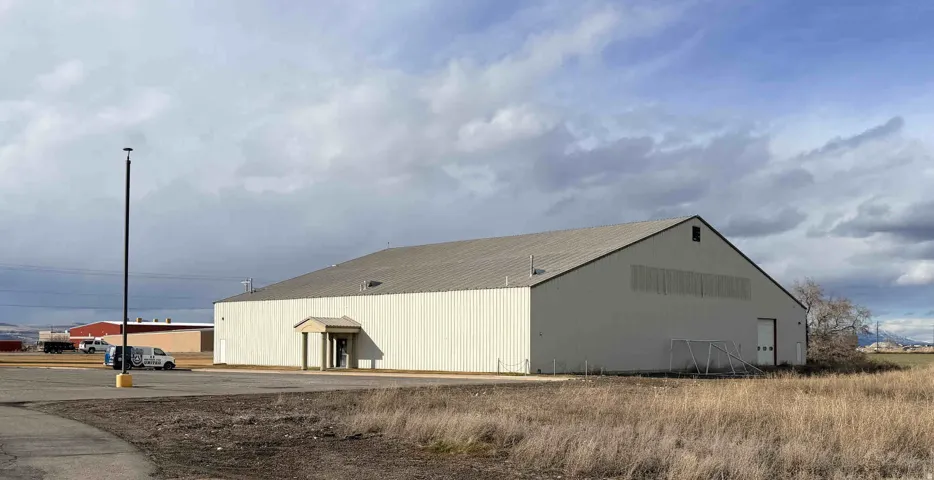 Metal-sided building with a pitched roof, featuring an entry awning, a large garage door, and ample paved parking