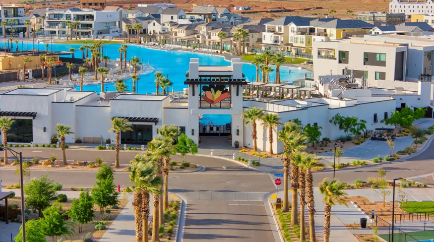 Community entrance featuring a central roadway, mature palm trees, contemporary white architecture, and a large recreational water feature