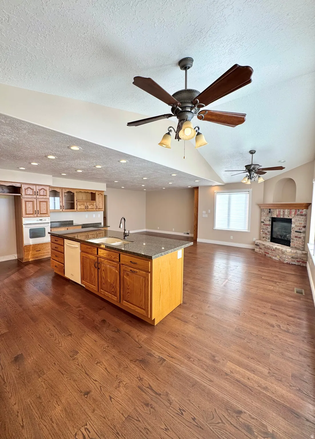 Open-concept living space featuring wood-finish flooring, a kitchen island with stone countertop, recessed lighting, and a brick-surround fireplace