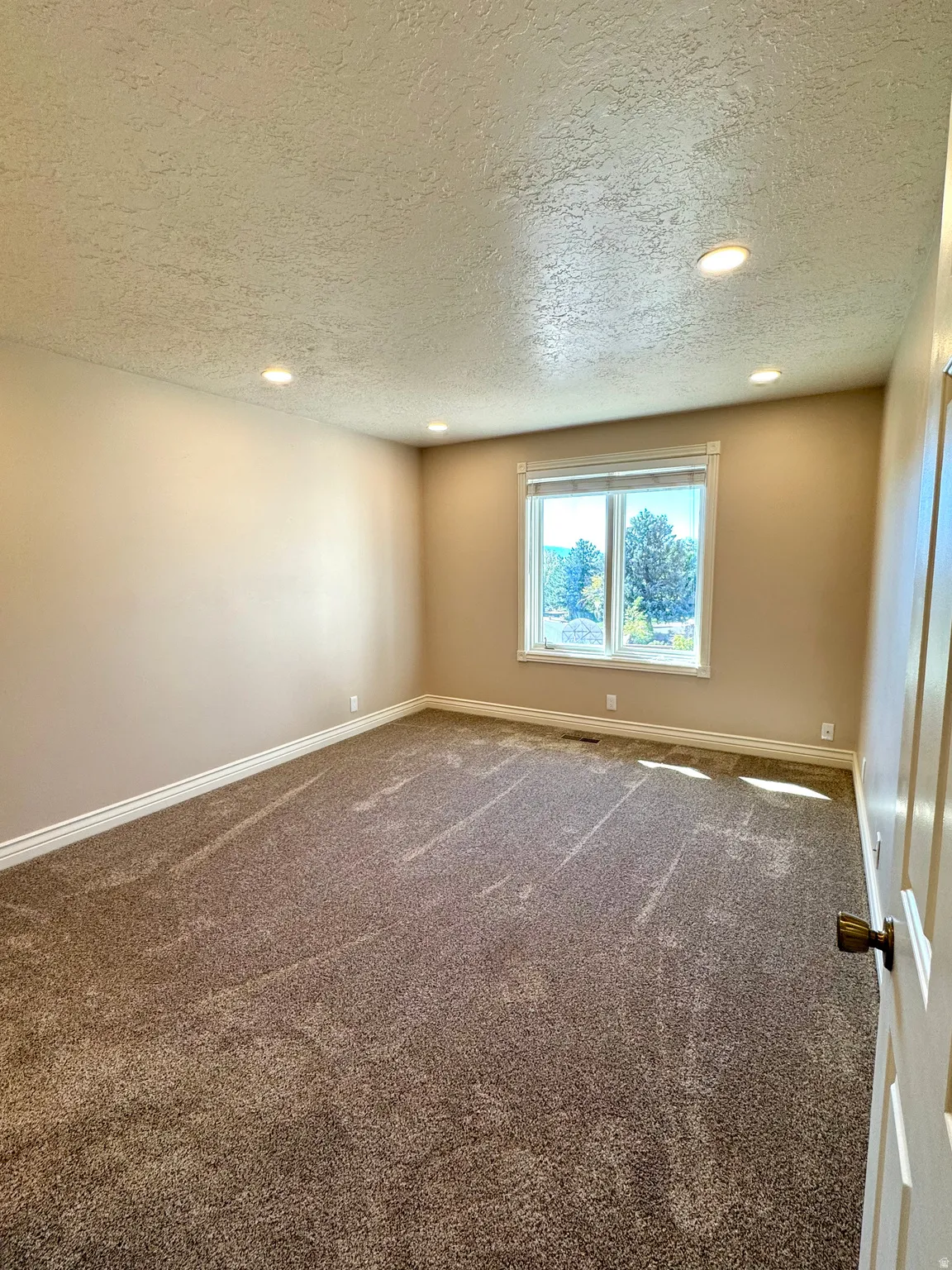 Recessed ceiling lighting, textured ceiling, light neutral wall paint, window with white trim, and brown carpet flooring