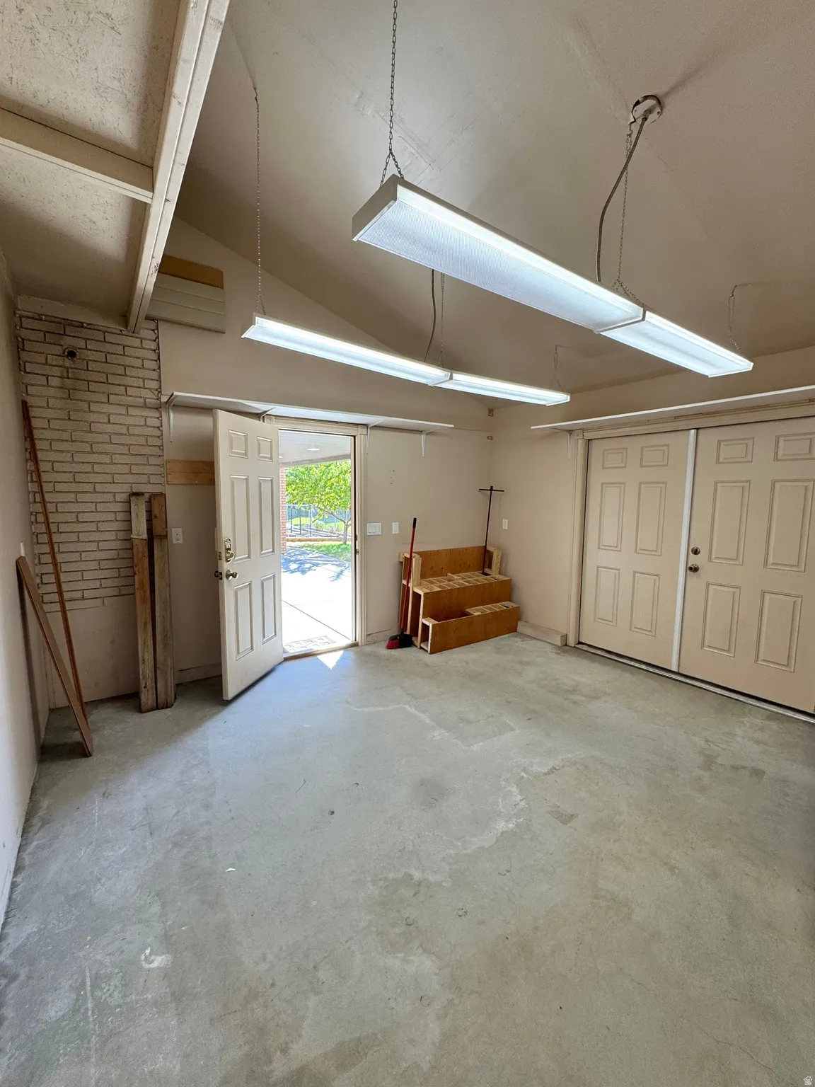 Vaulted ceiling with exposed beams, brick accent wall, concrete flooring, exterior access door, and double garage doors