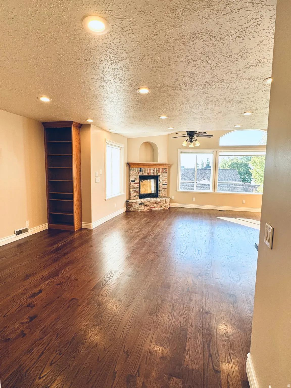 Spacious living area featuring rich wood-finish flooring, a stone-faced fireplace with a wood mantle, built-in shelving, a ceiling fan, and recessed lighting