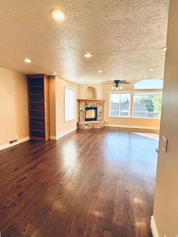 Spacious living area featuring rich wood-finish flooring, a stone-faced fireplace with a wood mantle, built-in shelving, a ceiling fan, and recessed lighting