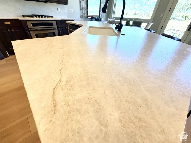 Kitchen island with integrated sink and matte black faucet, light-toned countertop with natural veining, stainless steel oven, dark wood cabinetry, and wood-finish flooring