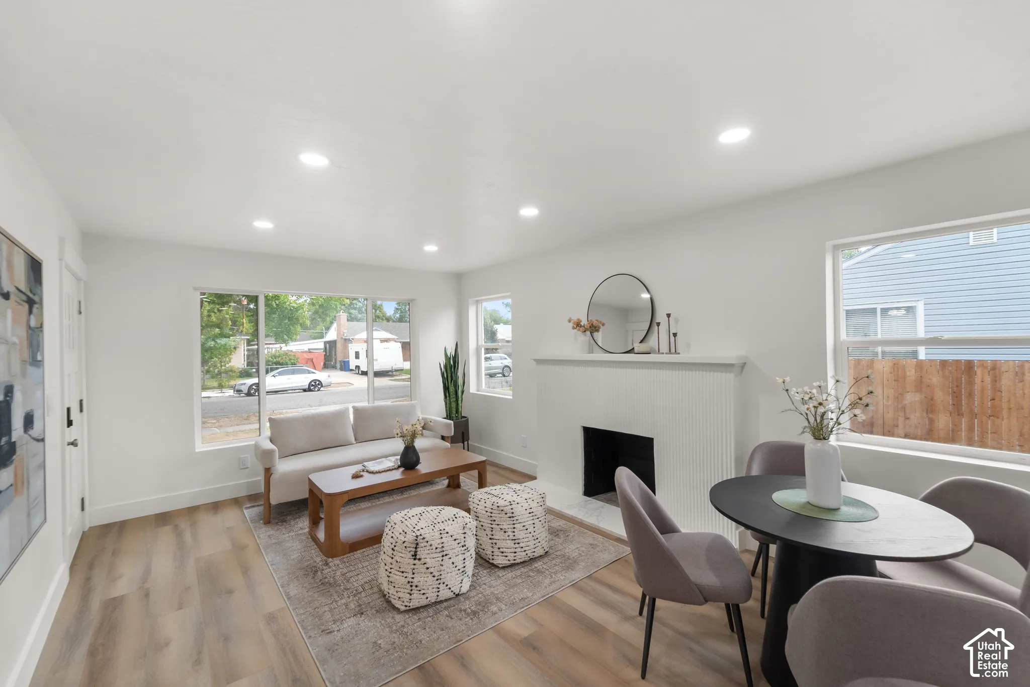 Recessed ceiling lighting, wood-finish flooring, white painted walls, white fireplace with hearth, and large windows