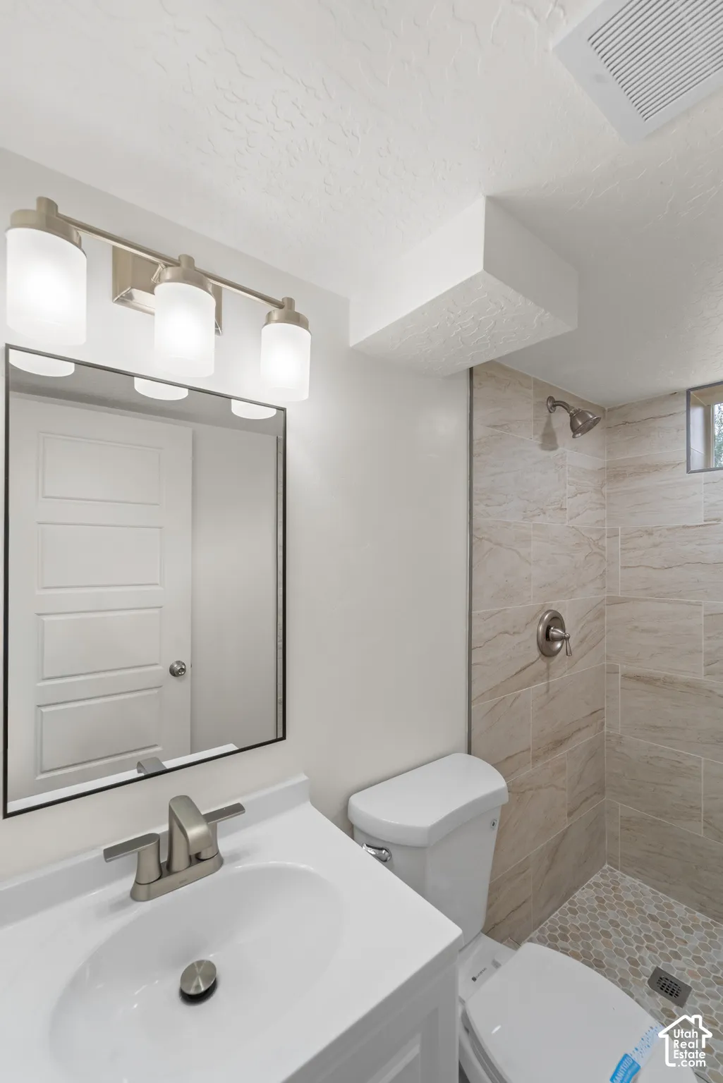 Vanity with an integrated sink and brushed nickel faucet, mirror with a black frame, three-light vanity fixture with frosted glass shades, shower with light-toned tile walls and a pebble-tiled floor